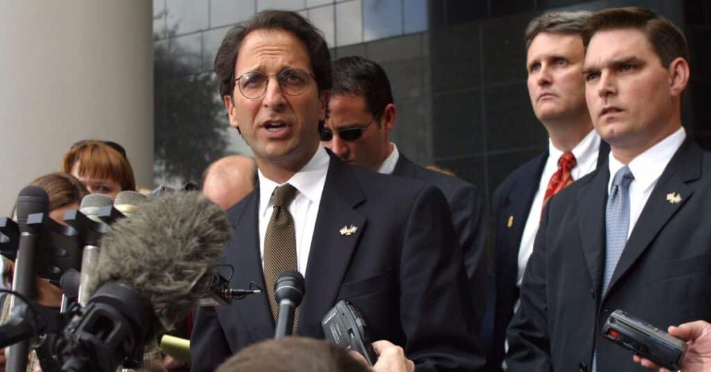 Surrounded by reporters, a man in a suit and tie addresses the media, highlighting the intensity of the moment.